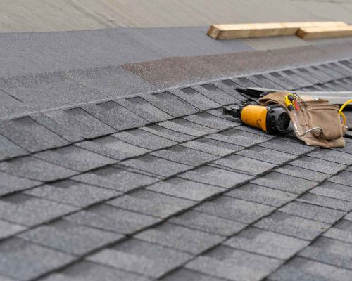 Photo of toolbelt with instrument and nail gun lying on asphalt or bitumen shingle on top of the new roof under construction residential house or building